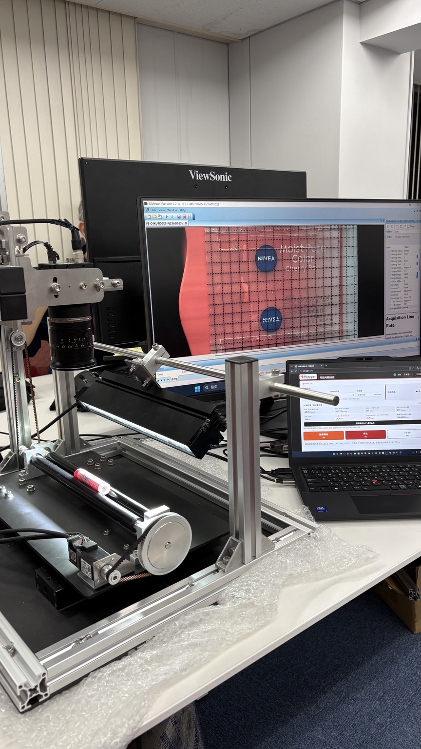 Full inspection setup with a pink lip balm tube on the rollers, the line-scan camera, operator interface, and captured surface image on the monitor